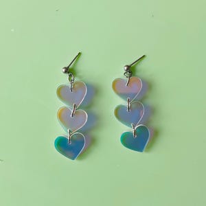 Puede incluir: Dos pares de pendientes colgantes iridiscentes en forma de corazón. Cada pendiente presenta tres corazones graduados suspendidos de un poste plateado. Los corazones cambian de color de amarillo a azul, creando un efecto brillante. El fondo es verde claro liso.