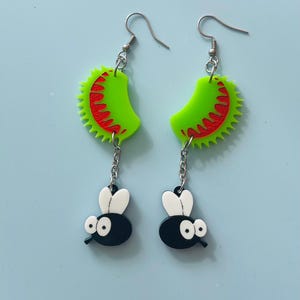 May include: Novelty earrings with a green Venus flytrap design, red detailing, and a black and white fly charm. The earrings have silver-coloured fish hook ear wires and chain links.
