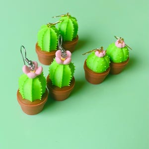 Peut inclure: Six boucles d'oreilles en forme de cactus vert avec des fleurs roses, chacune dans un petit pot marron. Les boucles d'oreilles ont des crochets argentés et sont posées sur un fond vert clair. Un accessoire amusant et fantaisiste.