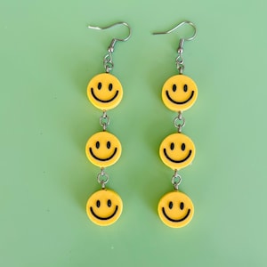 May include: Two pairs of dangling earrings, each featuring three yellow smiley faces with black eyes and mouths. The earrings are connected by silver chains and have silver fish hook closures. The background is a solid light green.