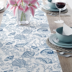 May include: A white table runner with a blue seashell and coral pattern. The runner is on a wooden table, with a vase of pink lilies, plates, bowls, and a wine glass visible.