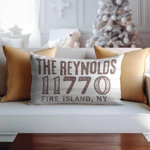 May include: A rectangular decorative pillow with the text "THE REYNOLDS 11770 FIRE ISLAND, NY" in brown lettering on a white and gray wood-grain background. Two gold-colored square pillows flank the decorative pillow on a white sofa.