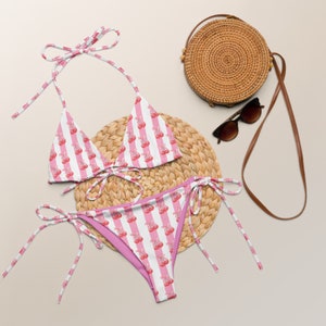 May include: Pink and white striped bikini with a cherry print. The bikini top is a triangle style with adjustable straps. The bikini bottom is a high-cut style with ties on the sides.