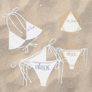 May include: White bikini sets with tie closures, featuring text in blue and gold. One set says "The future Mrs. Smythe" and another says "Bride babe". The other set says "Bride". All are on a sandy background.
