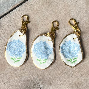 May include: Three keychains featuring white oyster shells with a gold trim and clasp. Each shell is hand-painted with a blue hydrangea flower design. The keychains are displayed on a light brown wooden surface.