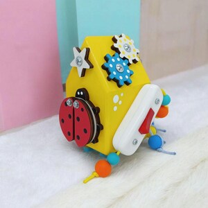 May include: A yellow wooden activity cube with various interactive elements. Features include a ladybug, gears, a star, a switch, and colorful beads. Designed for early childhood development and sensory exploration.