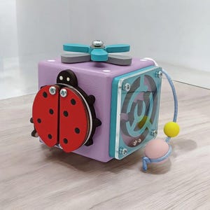 May include: A wooden activity cube with a ladybug, a flower, a maze, and a string with beads. The cube is painted purple, blue, and green.