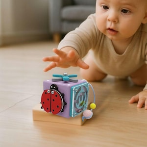 May include: A multi-activity wooden cube toy with a ladybug, maze, and spinning propeller. The cube is purple, red, and blue, designed for early childhood development. The toy sits on a wooden block.