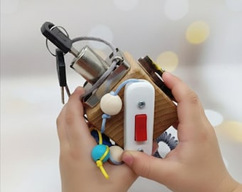 May include: A wooden activity cube featuring various interactive elements. It includes a key and lock, a light switch, and beads on a string. The cube is designed for sensory exploration and fine motor skill development.
