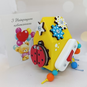 May include: A yellow wooden toy house with a ladybug, star, and gears. The house has a white door with a red button. The toy is attached to a string of colorful beads.