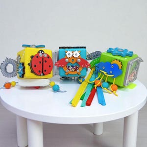 May include: Three colorful wooden activity cubes for toddlers. The cubes feature various interactive elements, including a ladybug, an owl, a cloud, and colorful ribbons. The toys are designed to stimulate sensory development and fine motor skills.