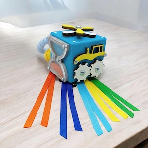 May include: A blue wooden activity cube for toddlers, featuring a tractor, gears, and a propeller. It has an hourglass, and colorful ribbons in orange, blue, yellow, and green. The cube is designed to stimulate sensory and fine motor skills.