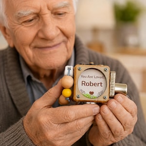 Op de afbeelding: Een houten fidget-kubus met een rond venster met de tekst "You Are Loved, Robert" en een klein bruin hartje. De kubus heeft een zijbevestiging met gladde houten kralen in natuurlijke en gele tinten, en een metalen zilverkleurige knop.