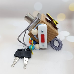 May include: A wooden activity cube with various sensory elements. Features a silver latch, a white switch with a red button, a gray cord with keys, and colorful beads. A gray coil and a yellow and brown shape are also attached.
