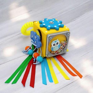 May include: A yellow wooden cube with a variety of textures and activities for babies. It features a spinning gear, a cloud with a rattle, a clear window with beads and an owl, and colorful ribbons.