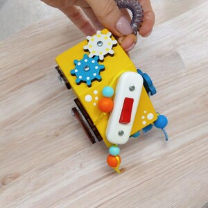 May include: A yellow wooden toy with colorful gears, a red button, and a string of beads. The toy is designed for babies and toddlers to explore and play with.