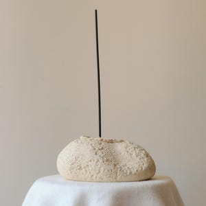 Peut inclure: Un porte-encens beige à la texture rocheuse et à la surface poreuse et irrégulière. Un bâton d'encens noir, fin et unique, est inséré verticalement au centre du support, s'étendant droit vers le haut depuis le sommet de la base en pierre.