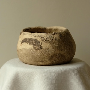 Op de afbeelding: Een kleine, ronde, handgevormde keramische kom met een getextureerd, onregelmatig oppervlak. De buitenkant heeft een gevlekt kleurenpalet van aardse beige, bruine en okerkleurige tinten, met een brede opening en een dikke, onregelmatige rand.
