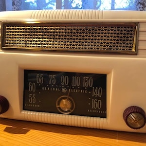 以下が含まれることがあります： 黒色の文字盤と金色のアクセントが付いた、白いビンテージのゼネラル・エレクトリックラジオ。文字盤には55から160までの数字と「GENERAL ELECTRIC」と「KILOCYCLES」の文字が書かれています。