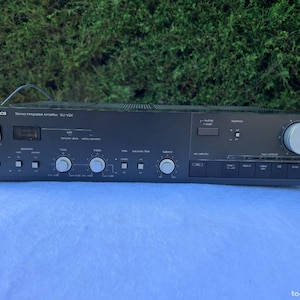 Puede incluir: Amplificador estéreo integrado Technics SU-V2X negro. El panel frontal tiene varias perillas de control, botones y una pantalla. El amplificador está sobre una superficie azul claro con un fondo verde.