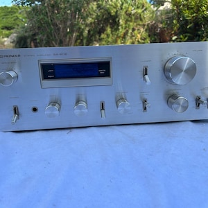 May include: Silver Pioneer SA-506 stereo amplifier with multiple knobs, switches, and a digital display. The amplifier has a brushed metal finish and is designed for audio equipment.