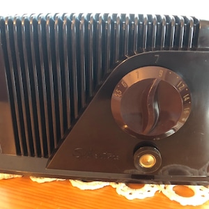 Puede incluir: Una radio de mesa vintage negra con un dial redondo y una perilla plateada. El dial tiene números del 5 al 16.