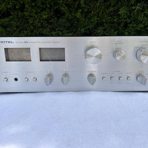Op de afbeelding: Zilverkleurige Rotel RA-714 stereo geïntegreerde versterker. De voorkant van de versterker heeft twee VU-meters, meerdere knoppen en knoppen voor volume, bas, treble en andere functies. De tekst "ROTEL STEREO INTEGRATED AMPLIFIER RA-714" staat bovenaan.
