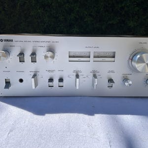 Puede incluir: Amplificador estéreo Yamaha CA-610 Natural Sound plateado. Cuenta con controles de volumen, graves, agudos y balance, además de varios selectores de entrada y salida. El amplificador tiene un botón de encendido, conector para auriculares y selector de altavoces.