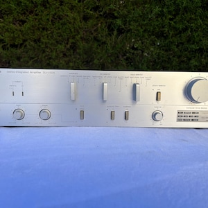 Op de afbeelding: Zilveren Technics Stereo Integrated Amplifier SU-V505 met diverse knoppen en draaiknoppen. De voorkant heeft tekst die de functies aangeeft. De versterker staat op een lichtblauwe ondergrond.