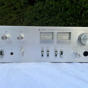 Könnte beinhalten: Silberner Sanyo Integrated Stereo Amplifier DCA 311. Die Vorderseite des Verstärkers hat Knöpfe für Leistung, Höhen, Bässe und Lautstärke. Es gibt auch Schalter für Lautsprecher, Hochfilter und Modus. Es gibt zwei Messgeräte und einen Kopfhöreranschluss.