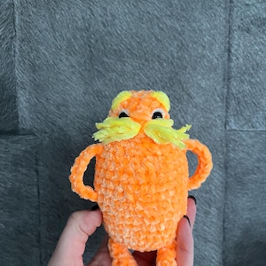 May include: Orange crocheted plush toy with yellow yarn for the moustache and black eyes. The toy is being held by a hand.