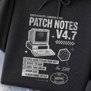 Könnte beinhalten: Ein dunkelgrauer Kapuzenpullover mit einem weißen Grafikdruck, der "Dungeon Master Campaign Update Patch Notes V4.7" liest, sowie einer Abbildung eines Computers, eines zwanzigseitigen Würfels und einer Liste von Spiel-Patch-Notizen.