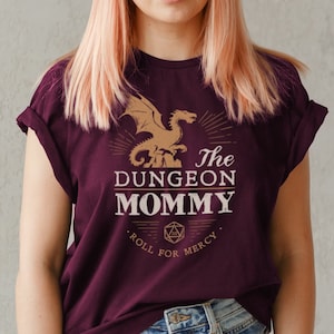 Può includere: Una maglietta color borgogna con un drago dorato e il testo "The Dungeon Mommy. Roll for Mercy."