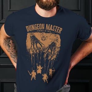 May include: Navy blue t-shirt with a brown graphic featuring the text "DUNGEON MASTER" and "STRINGS OF FATE". The graphic depicts hands manipulating puppet figures. A red beanie is worn.