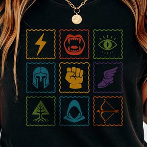 Könnte beinhalten: Schwarzes T-Shirt mit einem Raster aus farbenfrohen Symbolen. Die Symbole umfassen einen Blitz, einen Fangzahnmund, ein Auge, einen Helm, eine Faust, einen geflügelten Schuh, einen Baum, eine Kapuzenfigur und Pfeil und Bogen. Das Shirt ist ein Freizeitartikel.