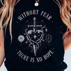 Könnte beinhalten: Ein schwarzes T-Shirt mit einem weißen Grafikdruck, der ein Schwert zeigt, das ein von dornigen Ranken umgebenes Herz durchbohrt. Der Text "WITHOUT FEAR" ist über dem Motiv gewölbt, "THERE IS NO HOPE" darunter. Zwei Spielwürfel sind zu sehen.