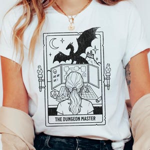 Może przedstawiać: Biały t-shirt z czarnym nadrukiem graficznym w stylu karty tarota. Ilustracja przedstawia osobę przy stole do gry ze smokiem, mapą lochów i kośćmi. Tekst na dole karty brzmi THE DUNGEON MASTER.