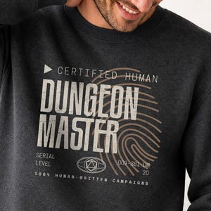 Pode incluir: Uma camisola de gola redonda em cinzento-carvão escuro com uma impressão gráfica que diz CERTIFIED HUMAN DUNGEON MASTER, um design de impressão digital, um ícone de dado de 20 lados e o texto 100% HUMAN-WRITTEN CAMPAIGNS em branco desgastado.