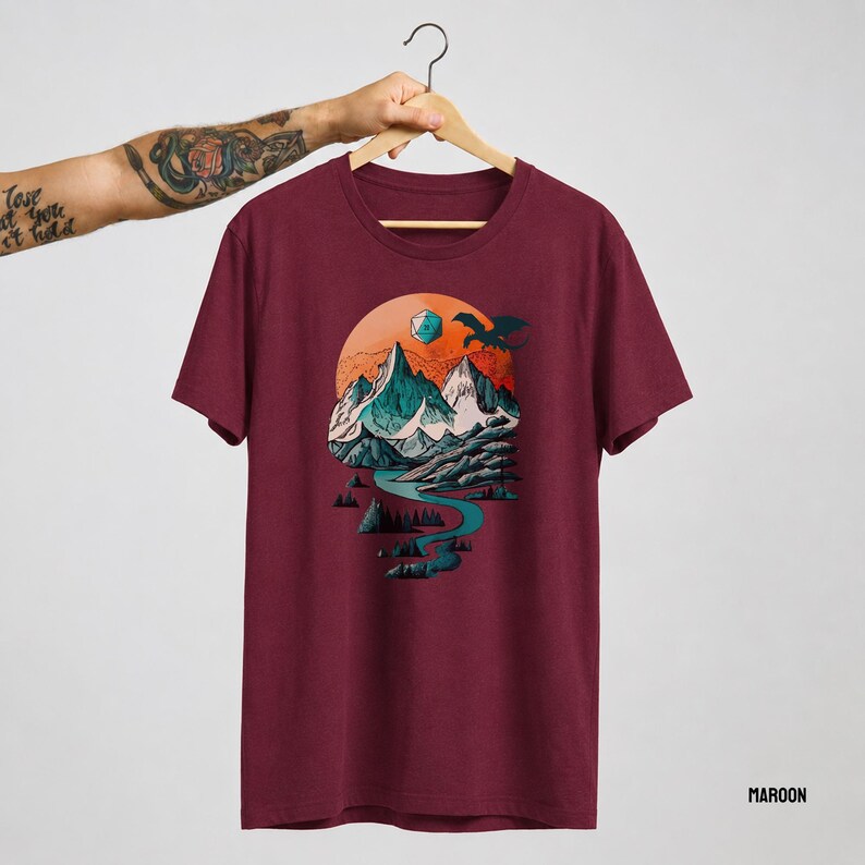 Maroon unisex t-shirt with the DnD adventure mountain design on a hanger — circular vintage landscape print featuring snowy peaks, pine trees, eagle, and D20 on a classic cotton tee