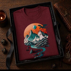 Maroon DnD mountain adventure shirt folded in a dark gift box — vintage circular landscape design as a fantasy gamer gift for tabletop RPG players and outdoor adventure fans