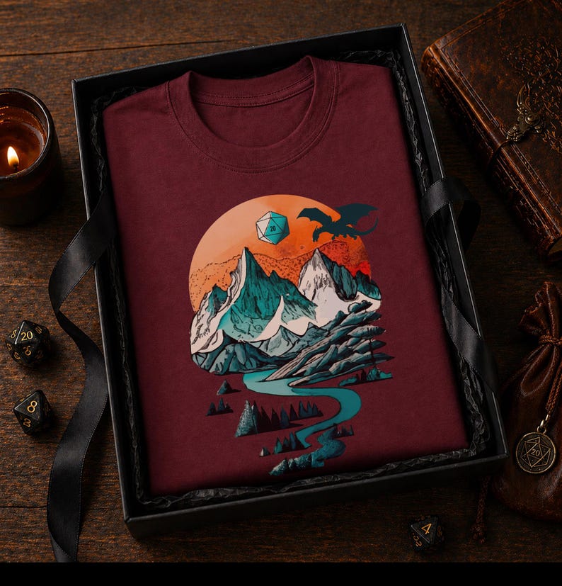 Maroon DnD mountain adventure shirt folded in a dark gift box — vintage circular landscape design as a fantasy gamer gift for tabletop RPG players and outdoor adventure fans