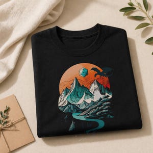 Folded black DnD mountain adventure shirt — front view showing the vintage circular landscape print with snowy peaks, D20, and eagle in teal and orange tones on cotton fabric