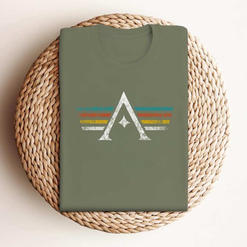 A minimalist t-shirt featuring the Arcs board game logo in a retro style. The design includes white geometric lines with a central star and vintage-colored stripes (teal, orange, red, yellow) on a dark background. Perfect for board gamers, sci-fi fans, and anyone who loves stylish and unique game night apparel.