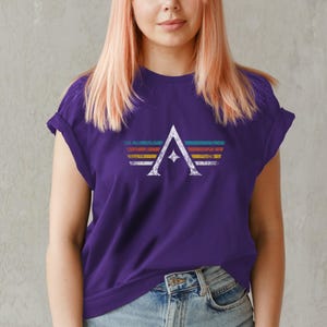 A minimalist t-shirt featuring the Arcs board game logo in a retro style. The design includes white geometric lines with a central star and vintage-colored stripes (teal, orange, red, yellow) on a dark background. Perfect for board gamers, sci-fi fans, and anyone who loves stylish and unique game night apparel.