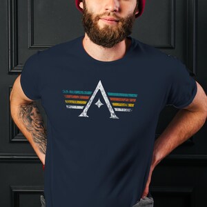 A minimalist t-shirt featuring the Arcs board game logo in a retro style. The design includes white geometric lines with a central star and vintage-colored stripes (teal, orange, red, yellow) on a dark background. Perfect for board gamers, sci-fi fans, and anyone who loves stylish and unique game night apparel.