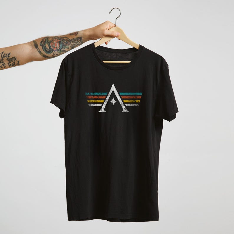 A minimalist t-shirt featuring the Arcs board game logo in a retro style. The design includes white geometric lines with a central star and vintage-colored stripes (teal, orange, red, yellow) on a dark background. Perfect for board gamers, sci-fi fans, and anyone who loves stylish and unique game night apparel.