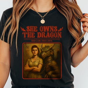 Può includere: T-shirt nera con la scritta rossa "SHE OWNS THE DRAGON" sopra l'immagine di una donna e un drago. L'immagine è in stile vintage. Il testo "HER LAIR. HER LAWS." è sotto.