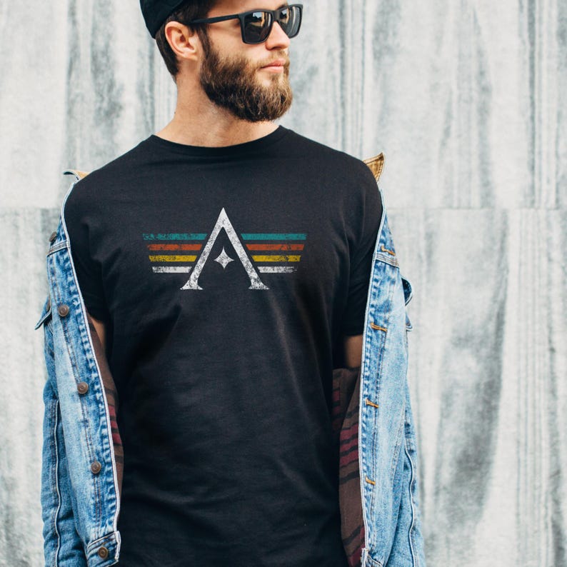 A minimalist t-shirt featuring the Arcs board game logo in a retro style. The design includes white geometric lines with a central star and vintage-colored stripes (teal, orange, red, yellow) on a dark background. Perfect for board gamers, sci-fi fans, and anyone who loves stylish and unique game night apparel.