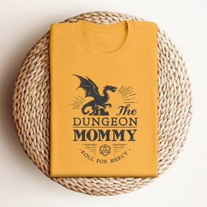 Dark red Dungeon Mommy T-shirt with a bold vintage-style dragon graphic above the phrase “Dungeon Mommy.” A large D20 dice and the words “Roll for Mercy” appear below, making this a funny and powerful gift for any Dungeons and Dragons fan. Ideal for gamer moms, dungeon masters, or fantasy lovers who want to show off their RPG pride in style. Perfect for birthdays, Mother’s Day, or any tabletop night.
