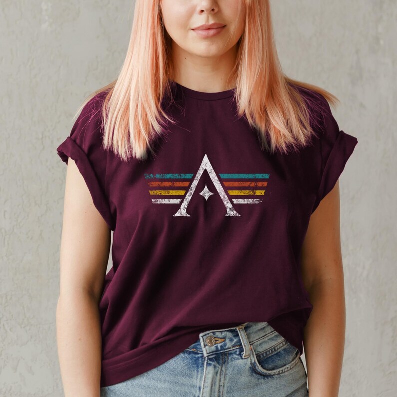 A minimalist t-shirt featuring the Arcs board game logo in a retro style. The design includes white geometric lines with a central star and vintage-colored stripes (teal, orange, red, yellow) on a dark background. Perfect for board gamers, sci-fi fans, and anyone who loves stylish and unique game night apparel.